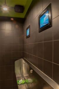 Baño de hombres con grandes azulejos oscuros, un urinario y pantallas de televisión montadas en la pared.
