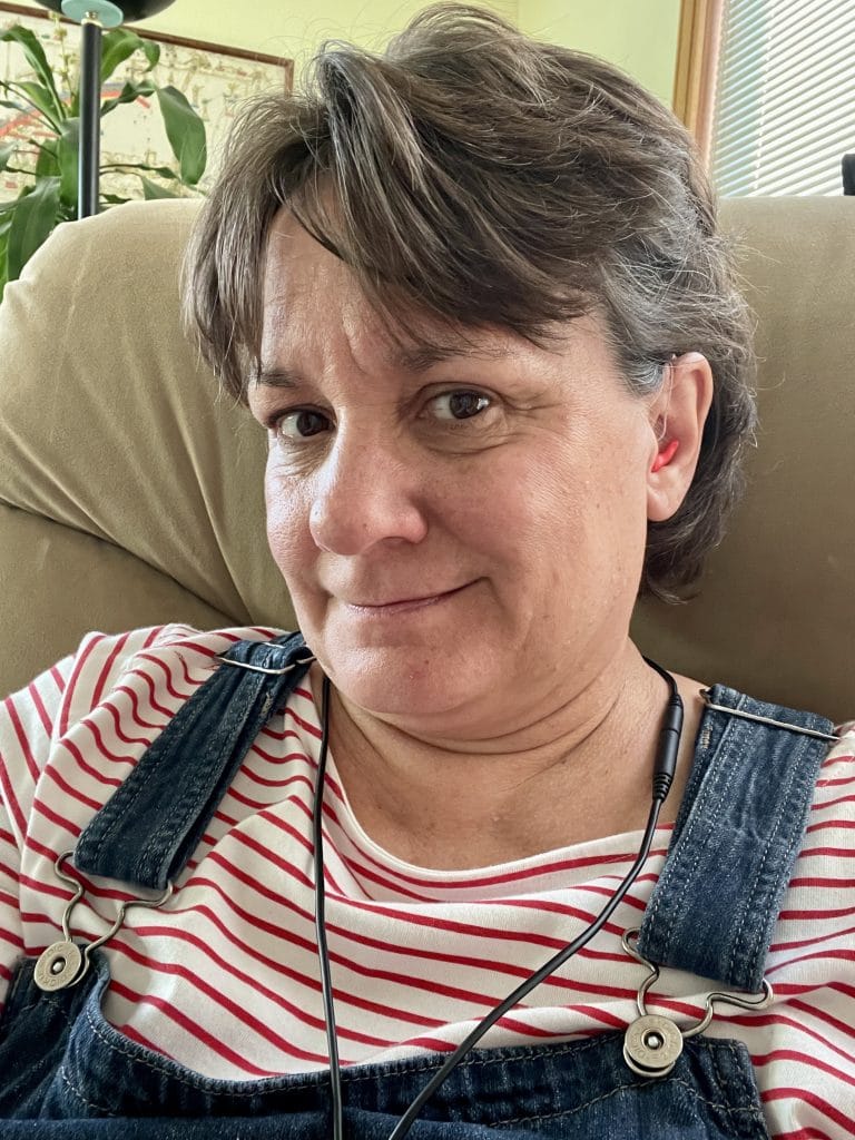 Cute picture of Chelle Wyatt sitting in her tan recliner wearing overalls, a red and white horizontally striped shirt, and the Listen Technologies advanced neck loop.
