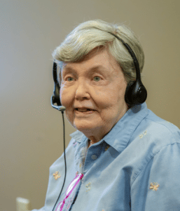 Crown Center resident, an older woman with short gray hair wearing a blue collared shirt wearing a ListenTALK headset and smiling.