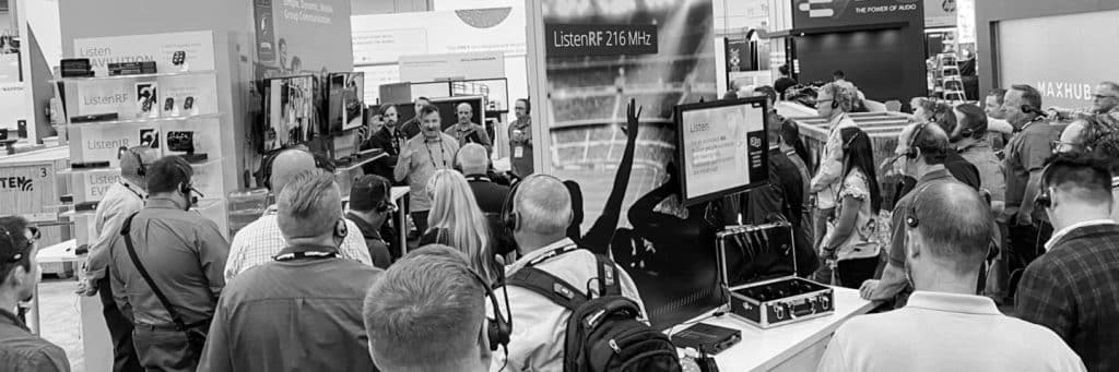 A tour group gathered at the Listen Technologies booth at InfoComm in 2022. Sam Nord is presenting and the group is wearing ListenTALK transceivers.