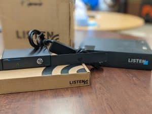 Small black box on top of Listen branded cardboard shipping box. The small server and box are sitting on top of a wood grain desk/surface.