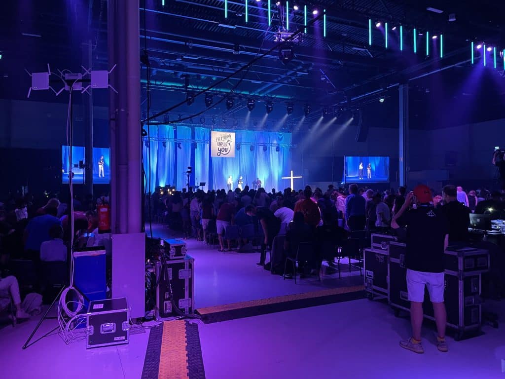 A view of the the event space filled with youth facing the front stage with theatrical lighting. Large TVs on both sides of the stage show the speakers on the stage.