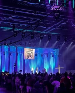 Una vista del espacio del evento lleno de jóvenes frente al escenario frontal con iluminación teatral. La parte posterior del escenario tiene largas cortinas azules y hay un letrero en el medio que dice: "Todo te desconecta", el tema principal. Cuatro personas están en el escenario junto con una cruz blanca a la derecha.