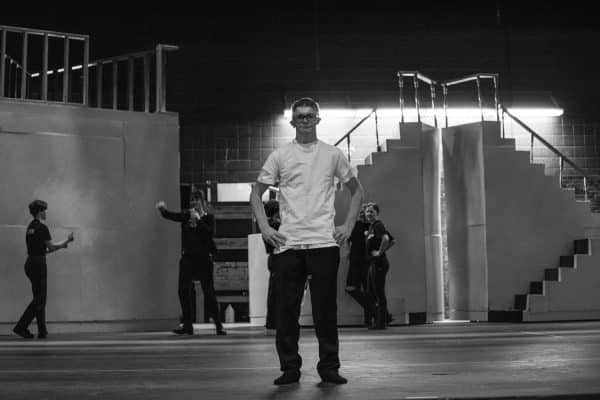 Black and white image of Fremont High Senior Hayden Reeve standing center stage wearing a white t-shirt and black pants with black framed glasses. The stage crew is in the background around the balcony stage props.