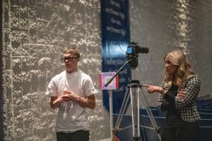 El estudiante Hayden Reeve y su maestro de artes escénicas y director de Anastasia, Aubrey Obray, parados en la parte trasera del teatro antes de una actuación. Hayden tiene el pelo corto de color fresa y viste pantalones negros y una camiseta blanca. La Sra. Obray tiene cabello rubio de longitud media y viste de negro con un blazer a cuadros de cintura corta.