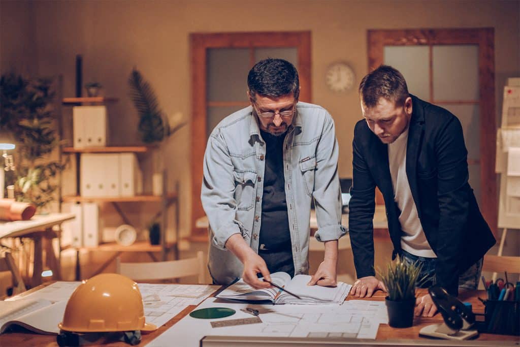 Two men standing over architectural drawings on desk with a hard hat resting at the top of the desk.