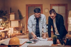 Dos hombres de pie sobre dibujos arquitectónicos en el escritorio con un casco descansando en la parte superior del escritorio.