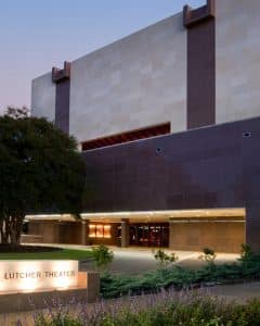 El exterior del Teatro Lutcher al anochecer en Orange, Texas. Hay un letrero iluminado que dice Lutcher Theatre, un gran árbol con un bonito paisaje. El edificio parece tener 4 o 5 pisos con materiales de construcción oscuros y claros alternados.