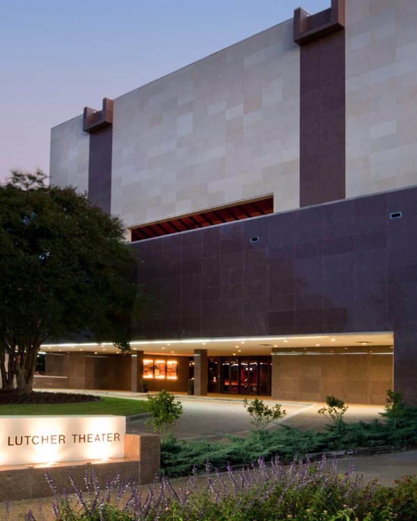 The outside of Lutcher Theater at dusk in Orange, Texas. There is a lighted sign that reads, Lutcher Theater, a large tree with nice landscaping. The building looks to be 4 or 5 stories with alternating dark and light building materials.