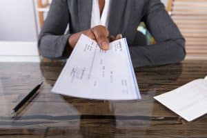 The torso of a business woman in a suit sitting at a desk handing a check across the desk.