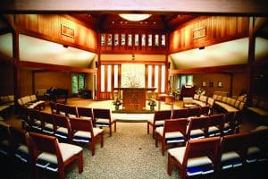 A photograph of the Farmington Valley Jewish Congregation - Emok Shalom sanctuary. Stained glass windows, with a tree are featured at the front of the sanctuary with a lecturn featuring the star of David. Rows of wide, padded, wood accent chairs are set up in a semi-circle around the lecturn.