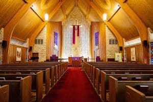 Beautiful A-frame sanctuary with wood paneled ceilings, brick walls, wooden pews, with red carpeting. A large back-lit cross with a maroon material artistically draped is featured behind the front lectern.