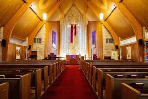 Magnifique sanctuaire en forme de A avec plafonds lambrissés, murs en briques, bancs en bois et moquette rouge. Une grande croix rétroéclairée avec un matériau marron artistiquement drapé est présentée derrière le pupitre avant.